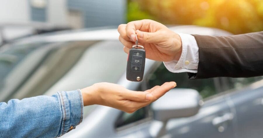 hand holding keys out to give to another hand; hand holding keys wearing business suit; hand accepting keys has a denim jacket on; silver car in background blurred